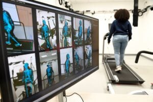 A monitor displays motion-capture skeletal visuals while a person walks on a treadmill in a lab setting.